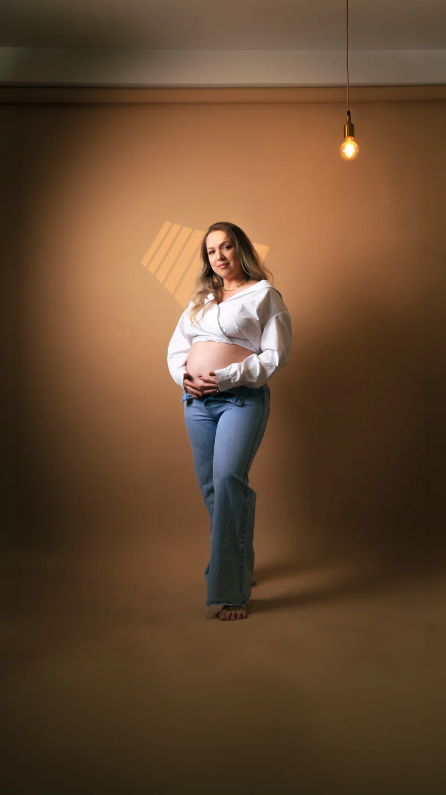 Deborah de corpo inteiro com camisa branca e jeans em fundo quente
