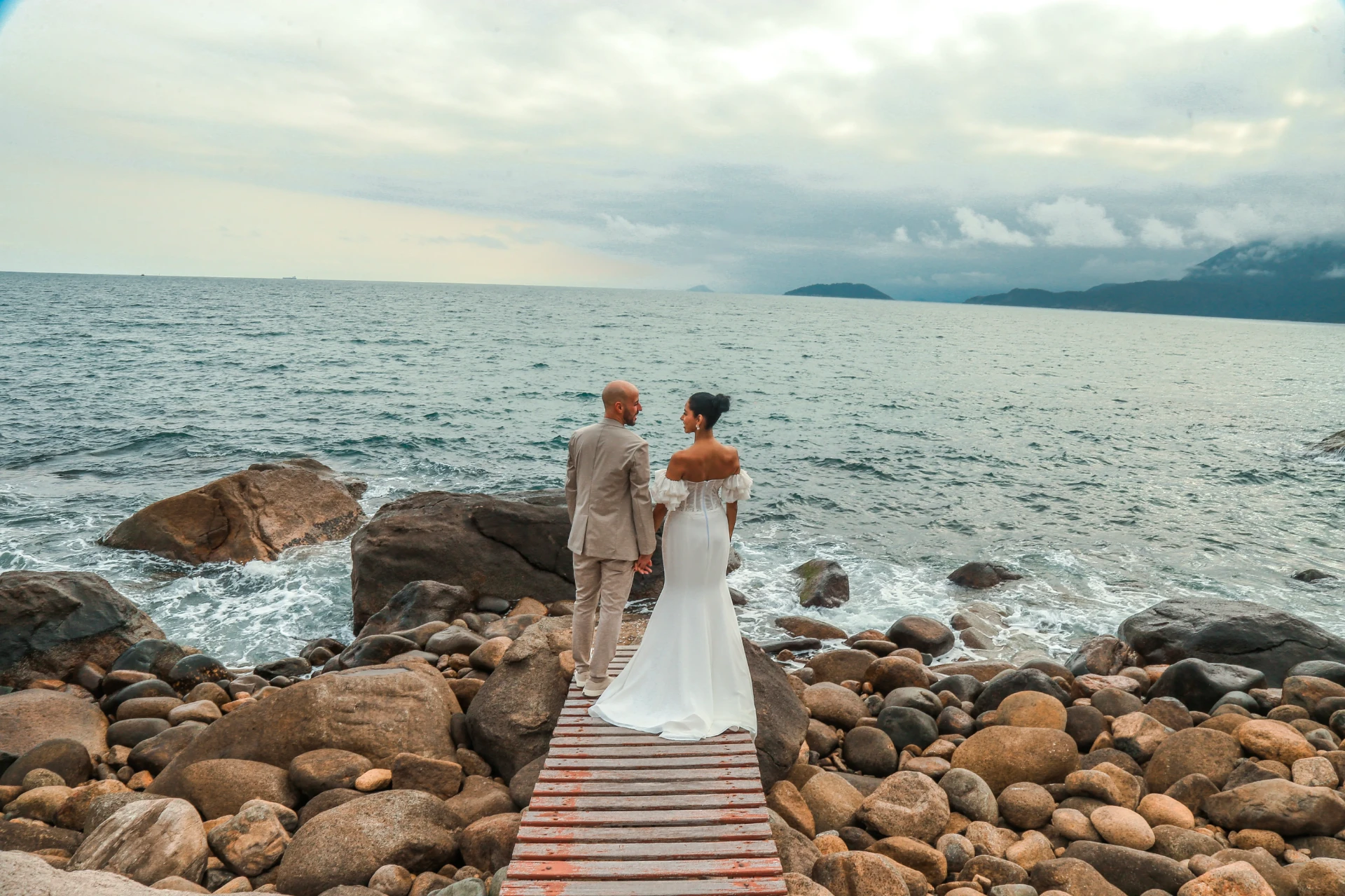 Larissa e Roberto — noivos de mãos dadas na passarela de madeira olhando o mar