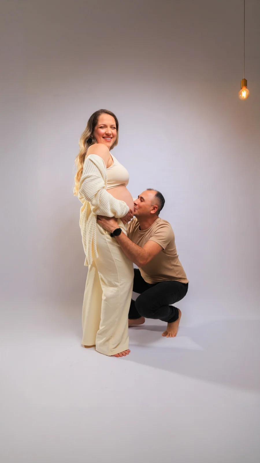 Wellington ajoelhado beijando a barriga de Deborah em fundo claro