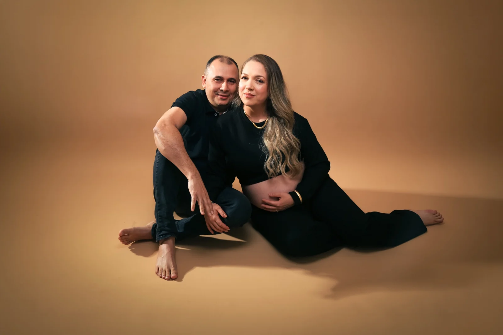 Deborah e Wellington sentados juntos de roupa preta em fundo bege